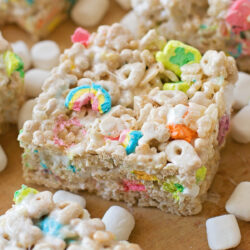 Cut Lucky Charm bars surrounded by mini marshmallows on a wooden board.
