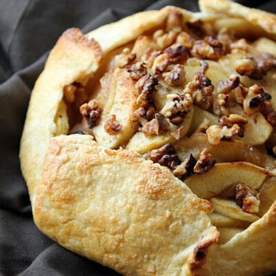 A rustic pear crostata with golden, flaky crust, topped with sliced pears and toasted walnuts, resting on a dark cloth.