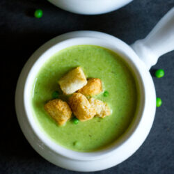 A bowl of green pea lentil soup, garnished with golden croutons, served in a white handled bowl.