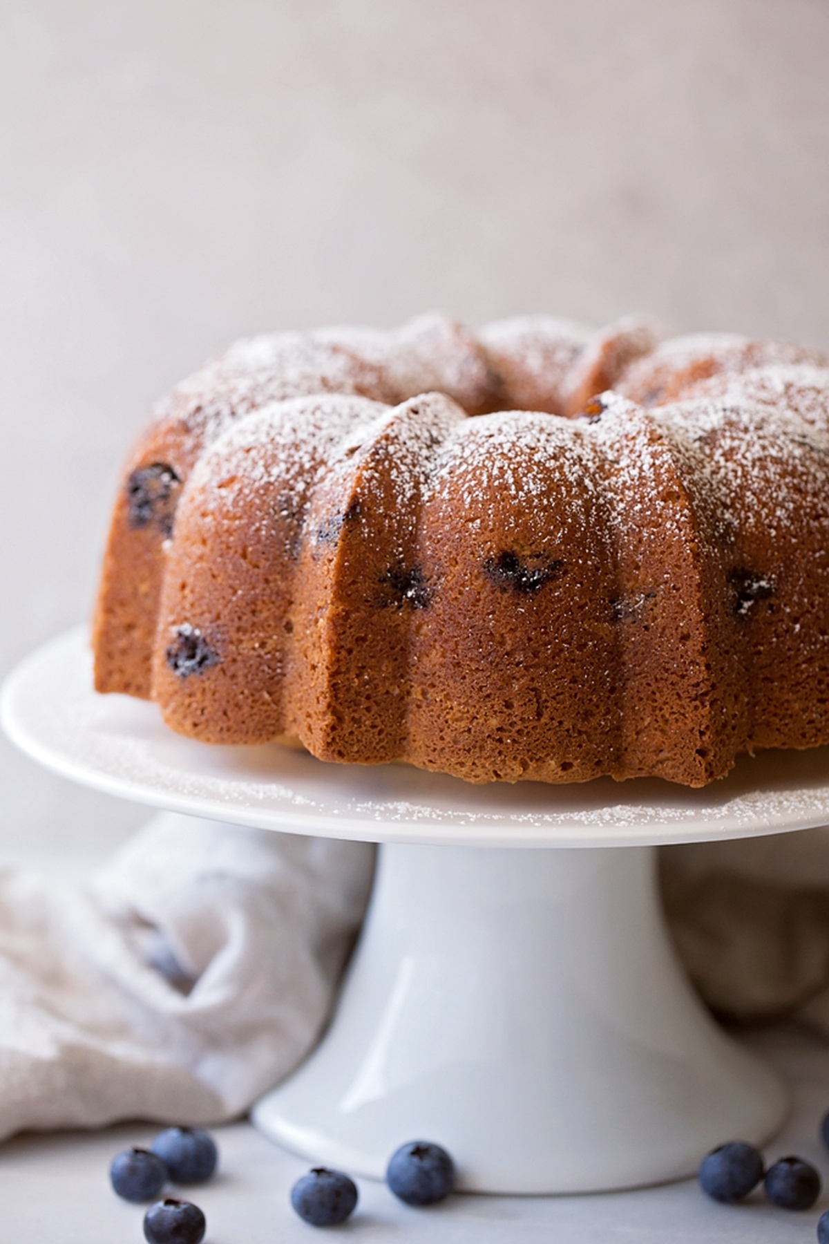 The whole Blueberry Almond Bundt Cake sitting on a cake stand, dusted with powdered sugar and surrounded by fresh blueberries.