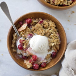 A wooden bowl filled with apple cranberry crisp, topped with a scoop of vanilla ice cream.