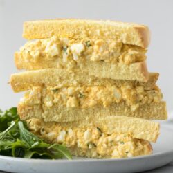 A stacked egg salad sandwich on soft white bread, served on a plate with fresh greens.