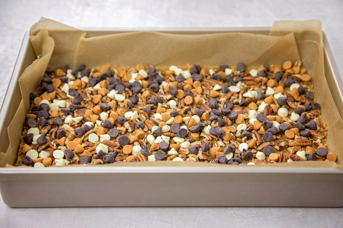 A baking pan lined with parchment paper, layered with chocolate chips, butterscotch chips, white chocolate chips, and pecans. 