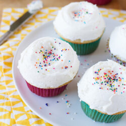 Vanilla bean cupcakes topped with icing for vanilla cupcakes and colorful sprinkles on a white plate.