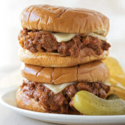 A stack of two Sloppy Joes with melted cheese, served with chips and a pickle.