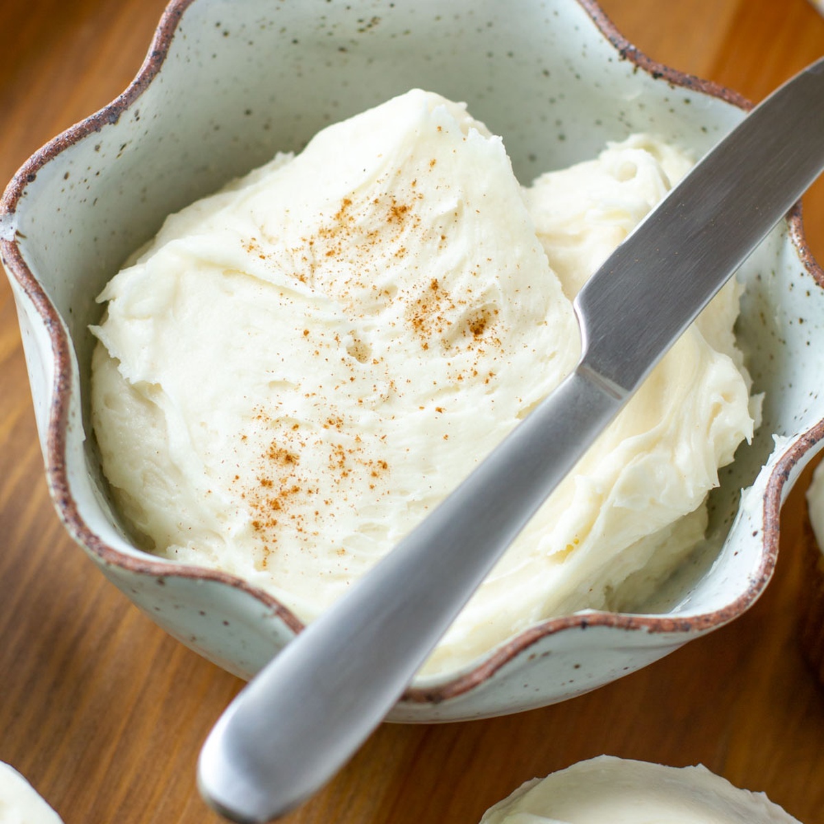 A bowl of creamy white cream cheese frosting topped with a sprinkle of cinnamon and a butter knife resting on the edge.