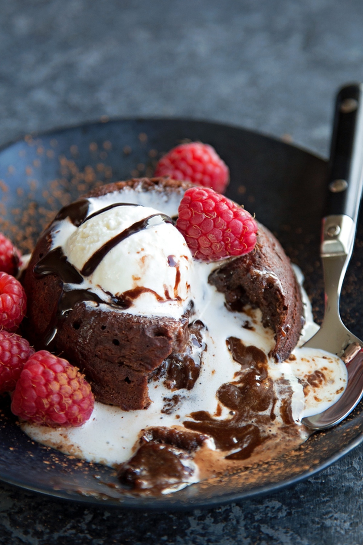 A rich chocolate molten lava cake topped with melting vanilla ice cream, drizzled with chocolate syrup, and garnished with fresh raspberries.