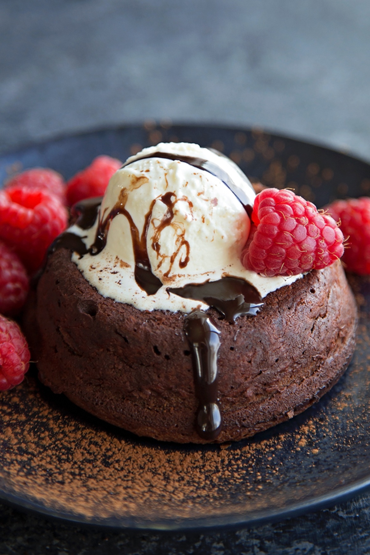 An individual chocolate lava cake topped with vanilla ice cream, chocolate drizzle, and raspberries, with cocoa powder sprinkled on the plate.