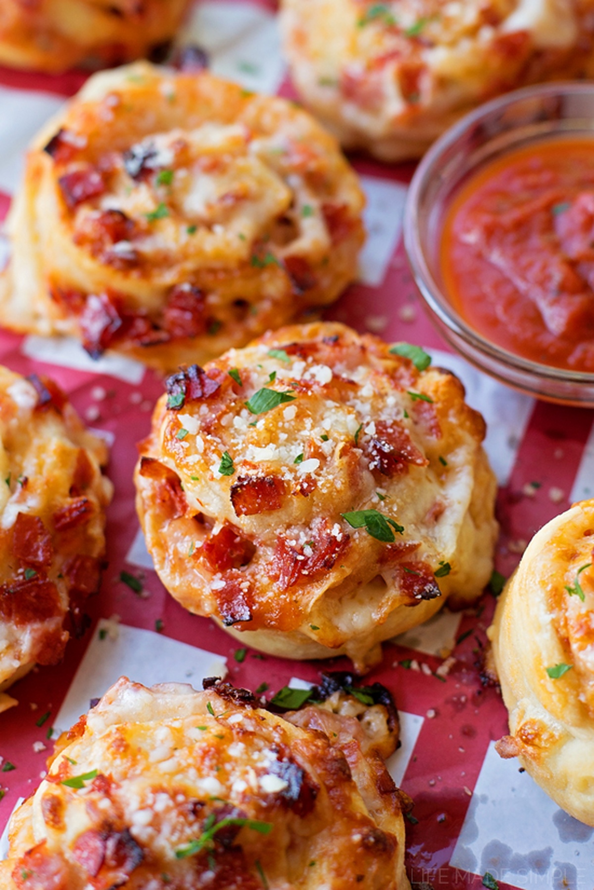These individual pepperoni pizza pinwheels are fun to make and eat! They're stuffed full of gooey mozzarella cheese and lots of crispy pepperoni. Several pepperoni pizza pinwheels arranged on red and white paper, with a small bowl of marinara sauce in the background.