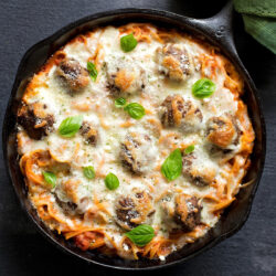 Spaghetti and meatballs bake topped with golden melted cheese and fresh basil leaves in a skillet.