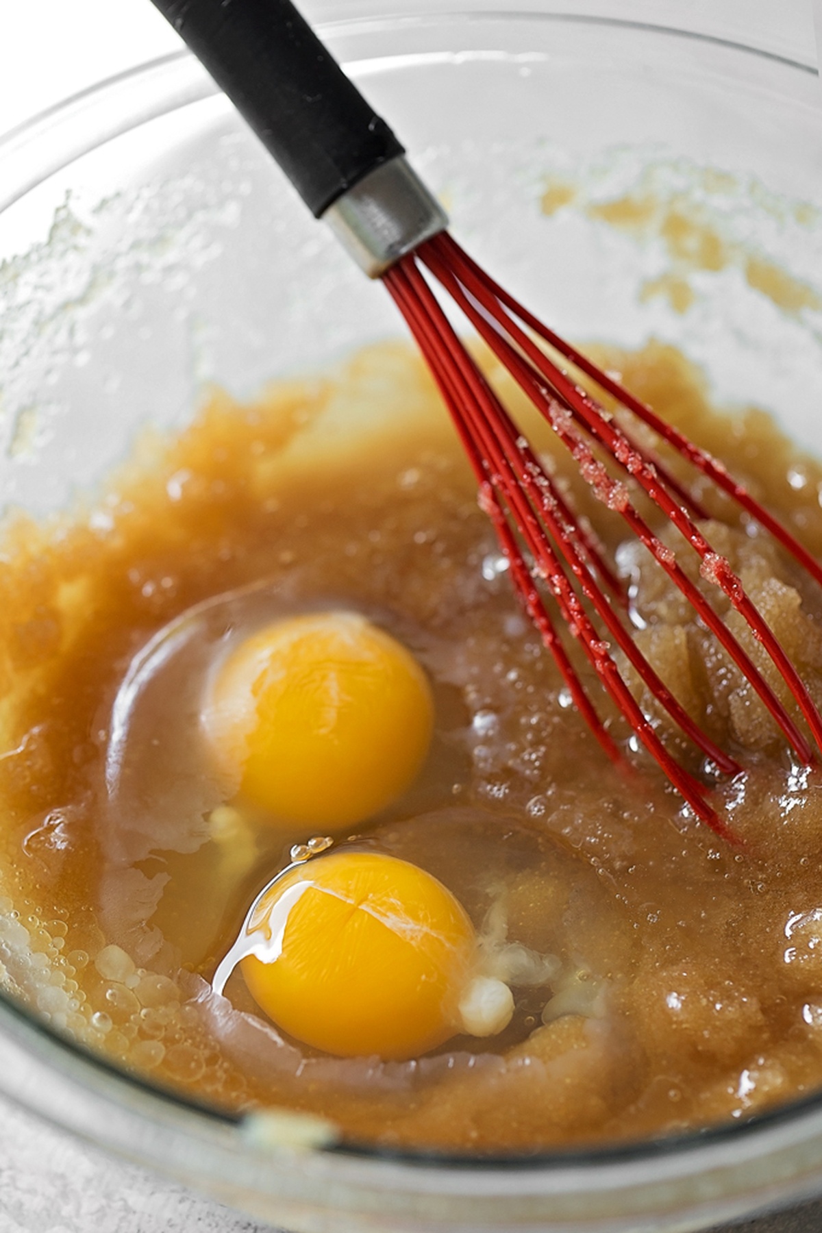 A glass bowl with a wet mixture of brown sugar and eggs being whisked using a red whisk.