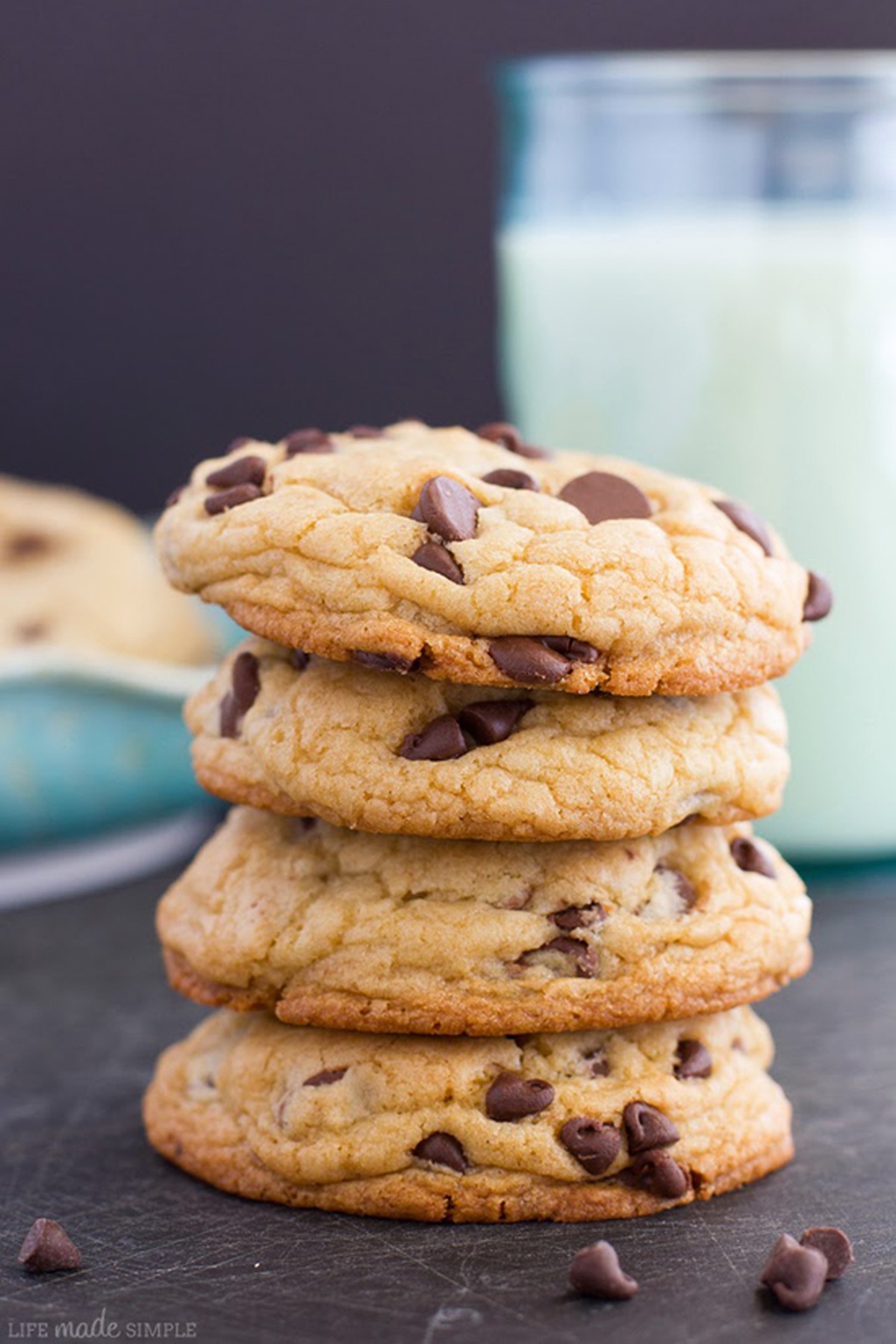 A stack of soft chocolate chip cookies on a dark surface with a glass of milk in the background.