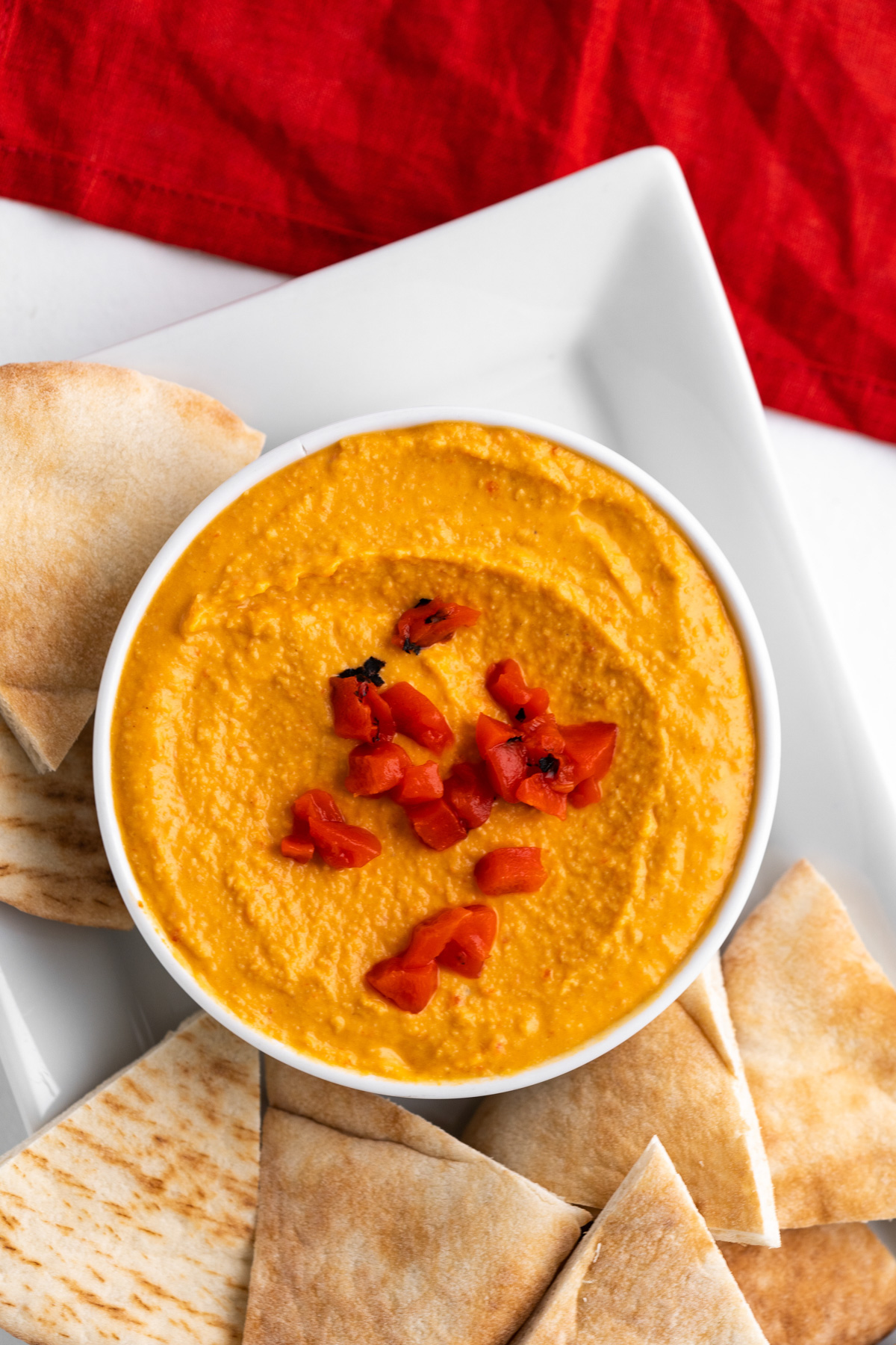 A bowl of red pepper hummus with pita bread triangles all around.