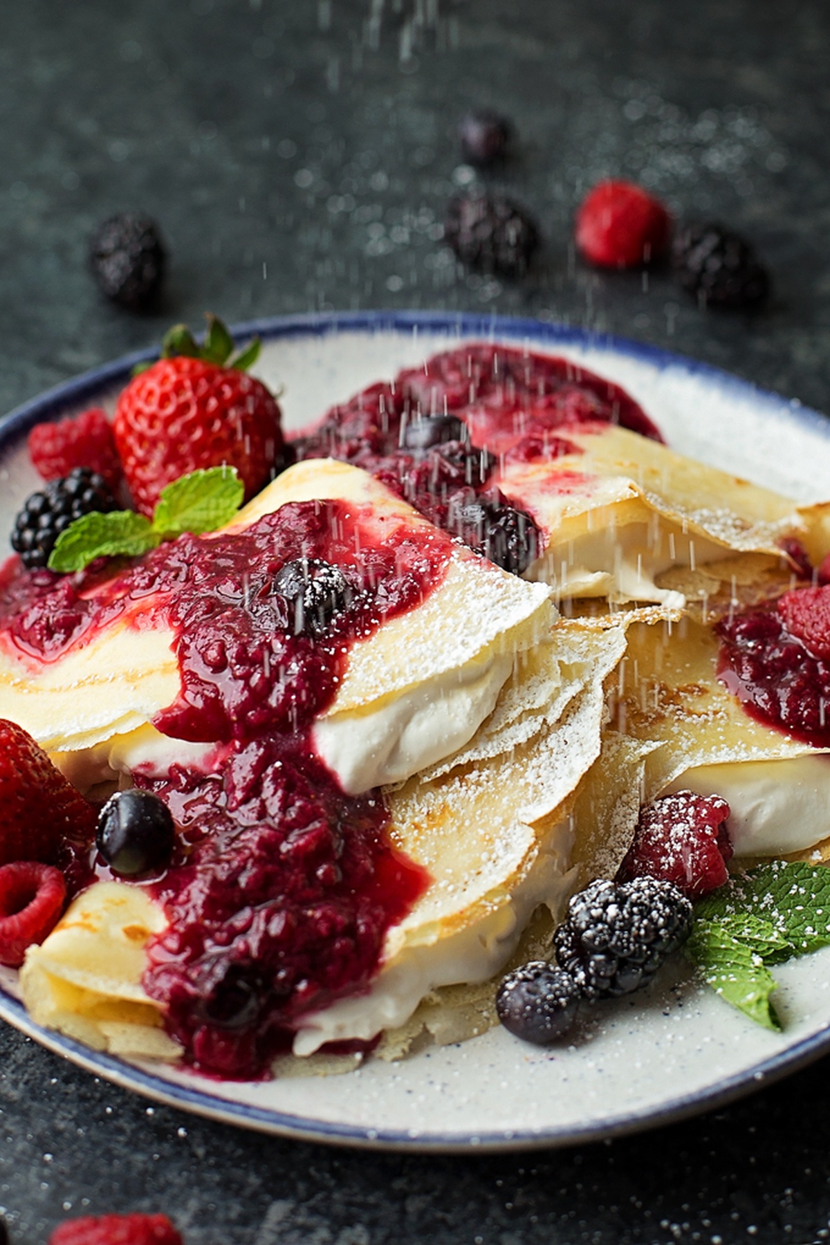 Sprinkling powdered sugar over fruit crepes with berry sauce.