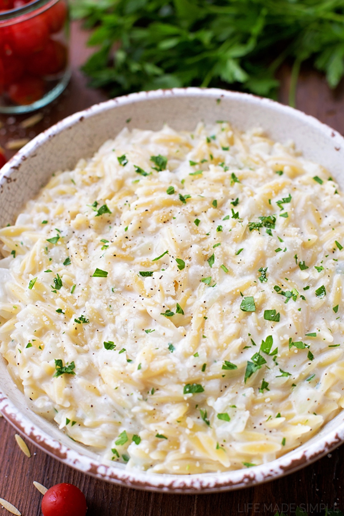 A serving dish filled with creamy Parmesan orzo, sprinkled with chopped parsley and black pepper.