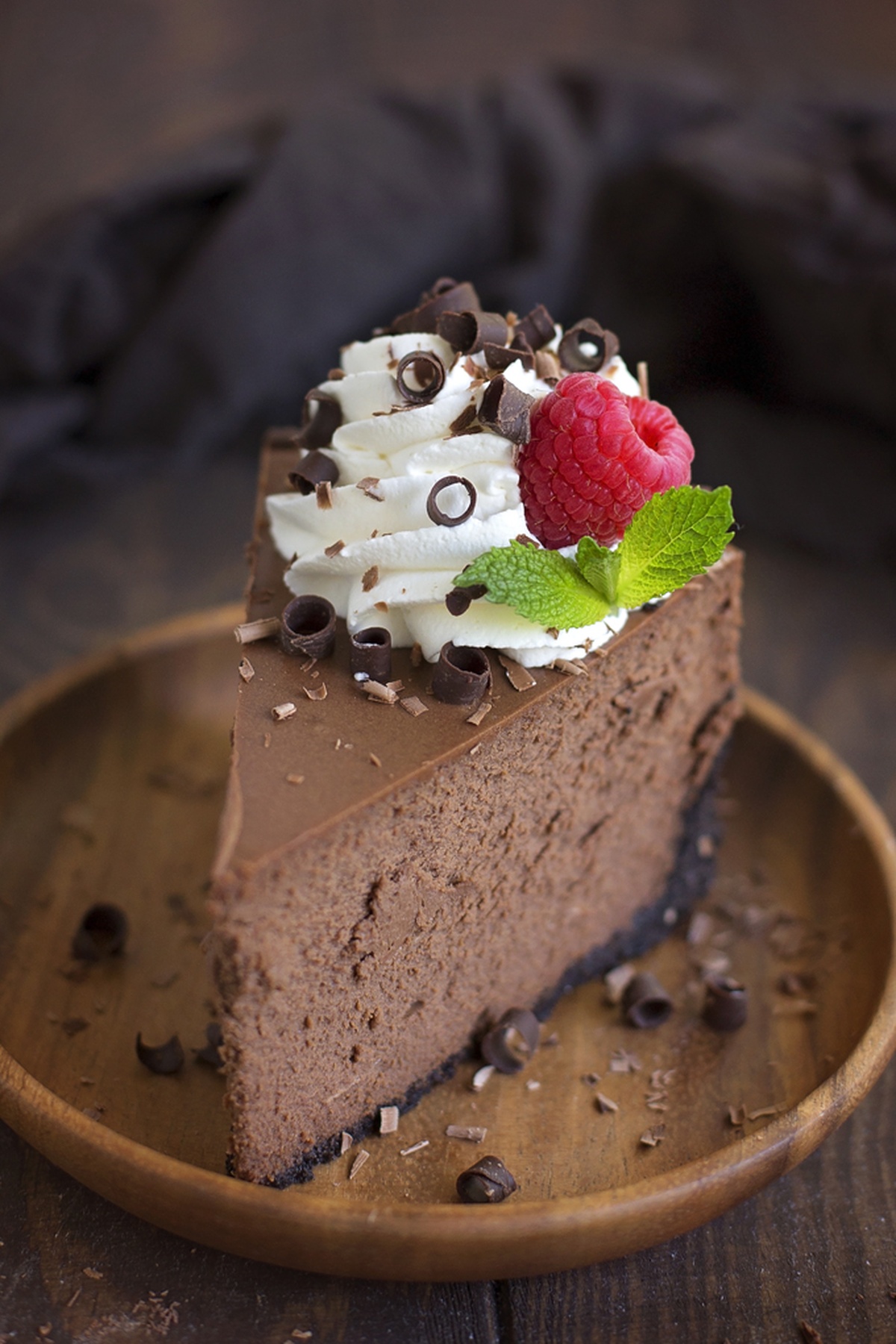 A single slice of chocolate cheesecake topped with whipped cream, chocolate curls, a raspberry, and mint on a wooden plate.