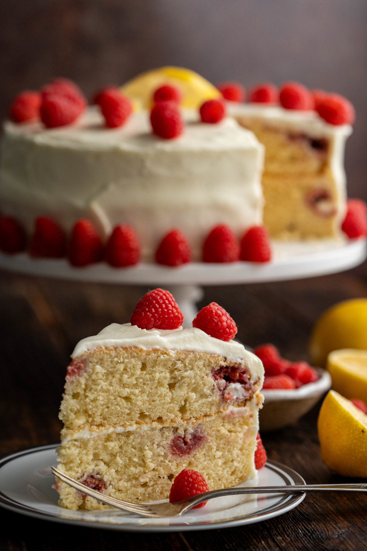 Slice of raspberry lemon cake on a white plate with the rest of the cake in the background.