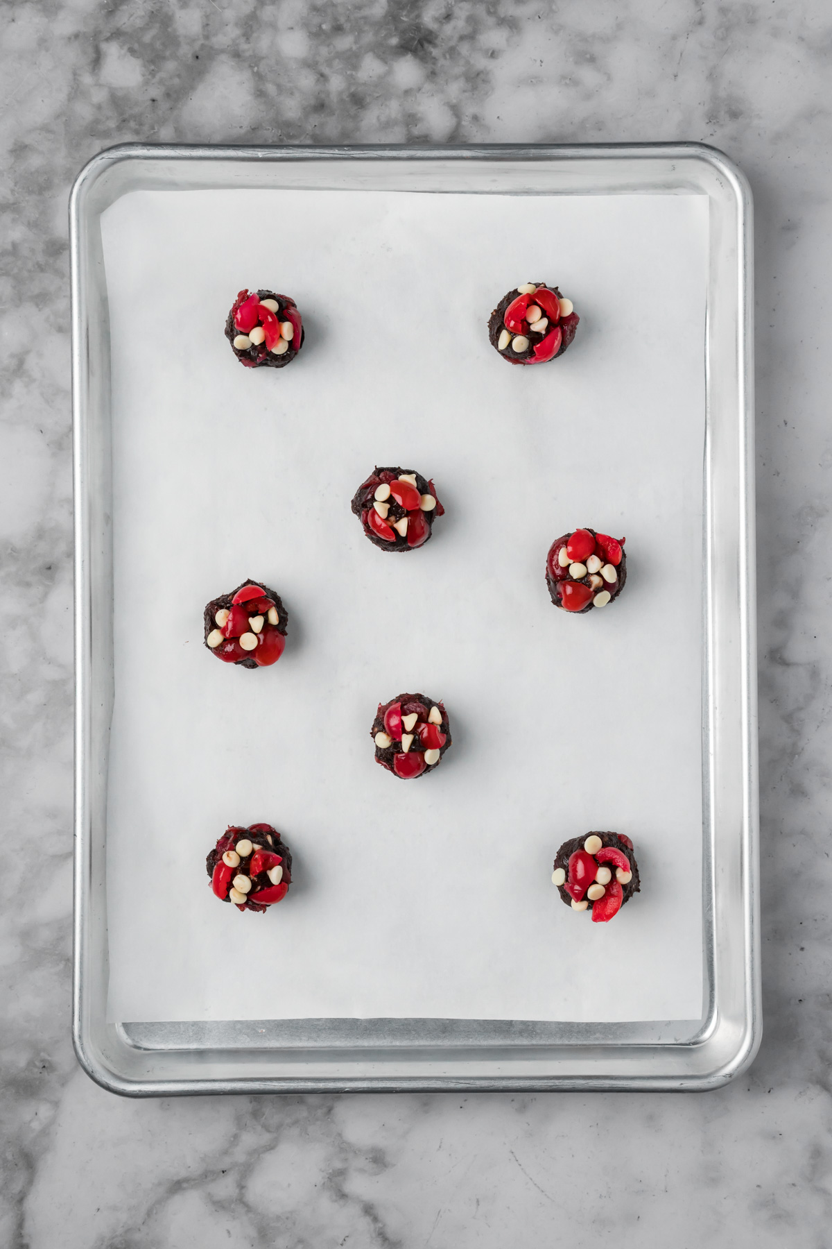 Cookie dough balls on a baking sheet with extra cherries and white chocolate chips on top.
